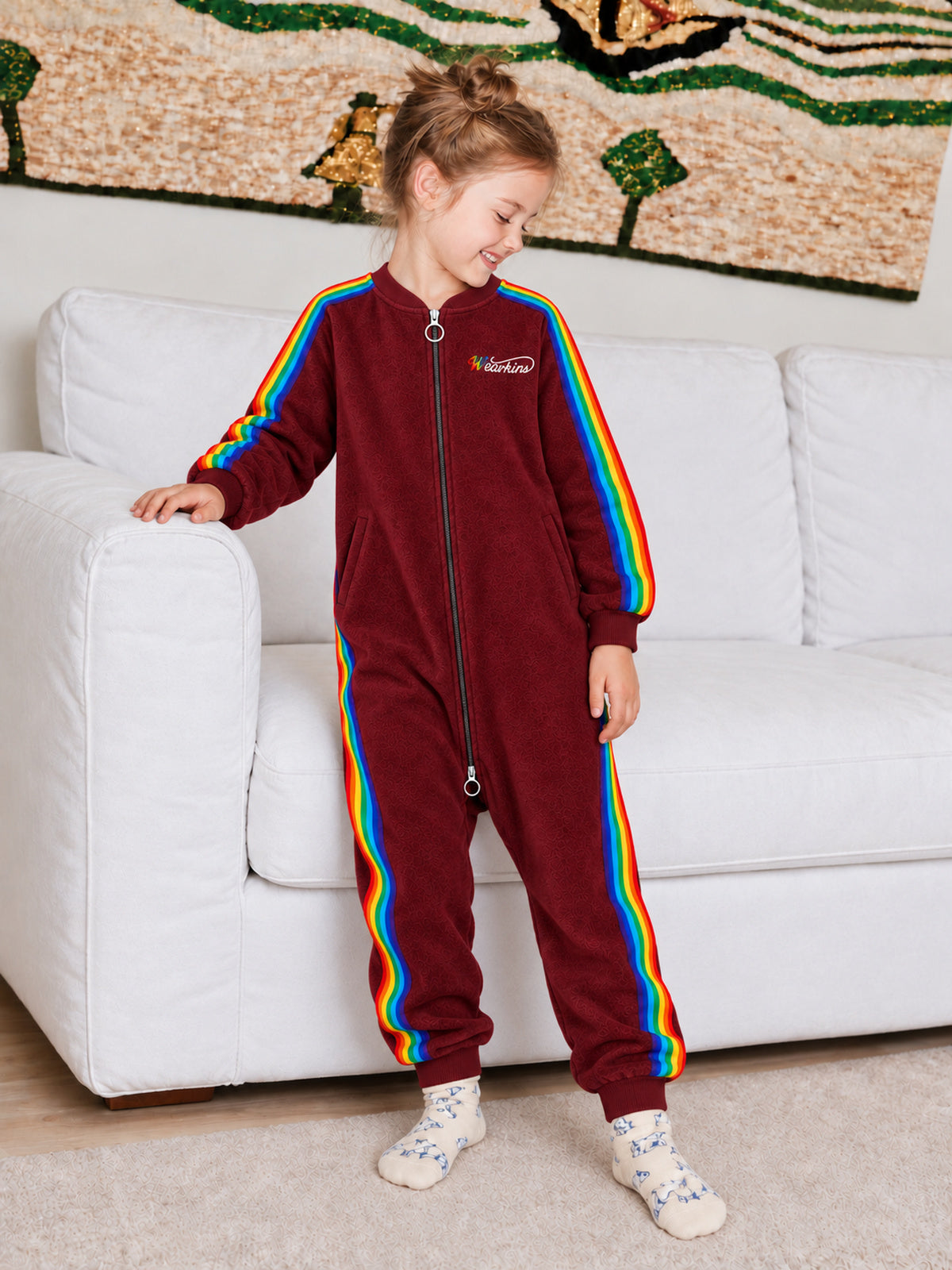 Child wearing a red rainbow-stripe onesie beside a sofa