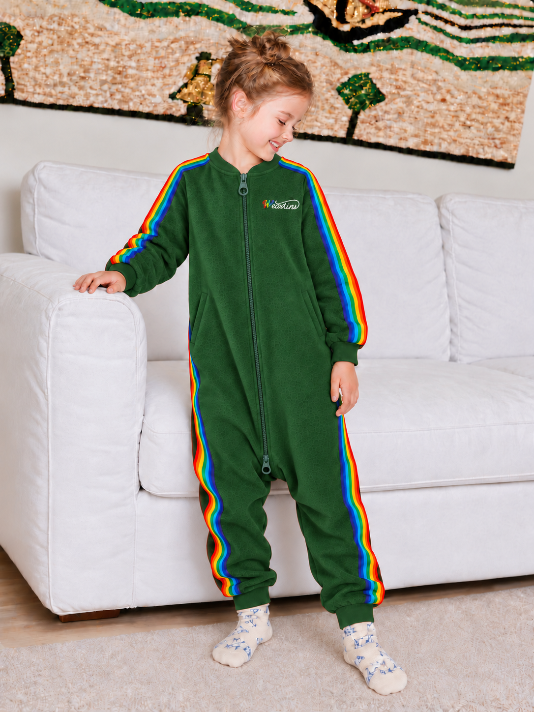 Child wearing a green rainbow-stripe onesie beside a sofa