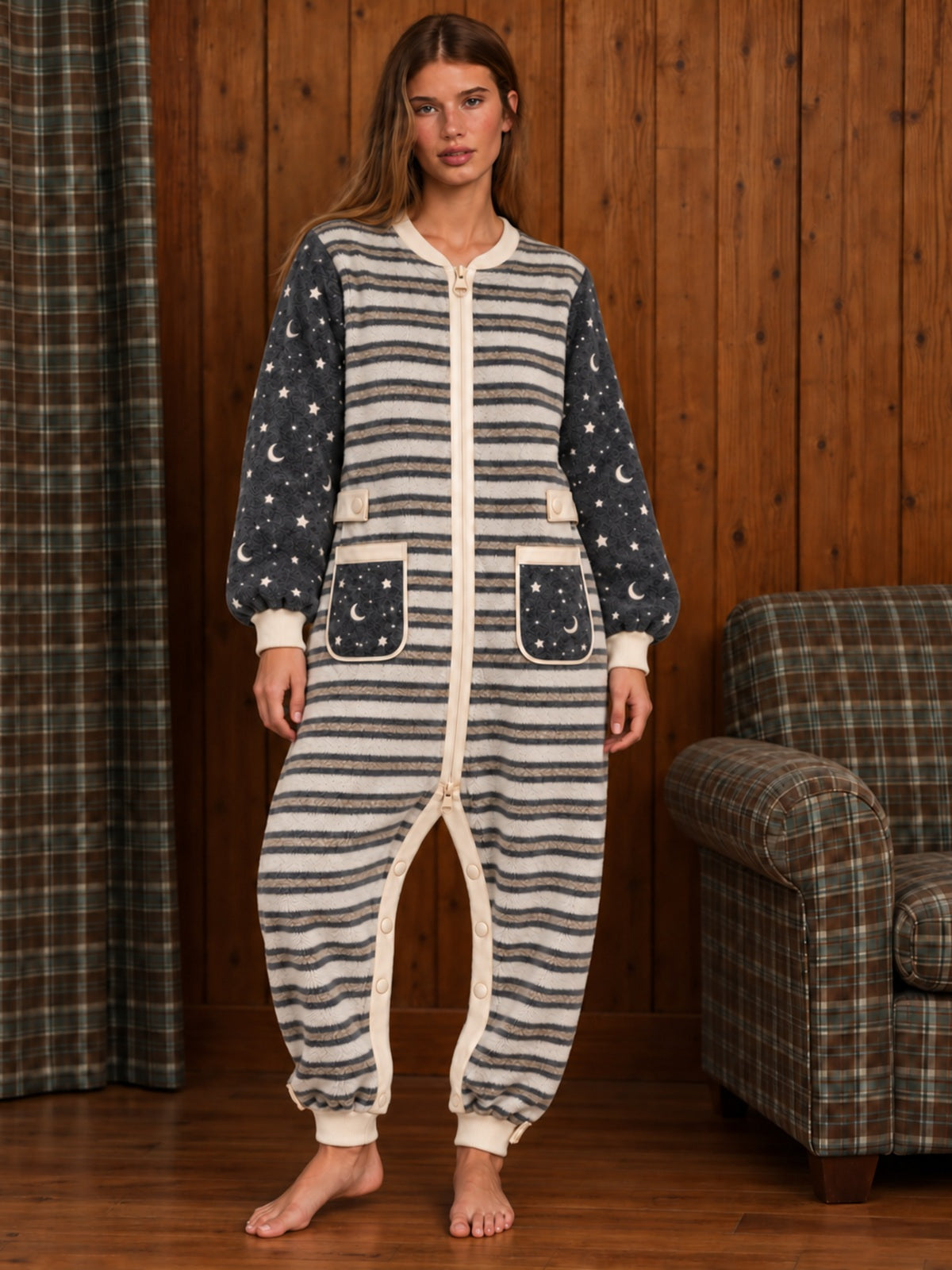 Adult female model wearing the star and moon striped onesie, standing in a wood-paneled room beside a plaid chair
